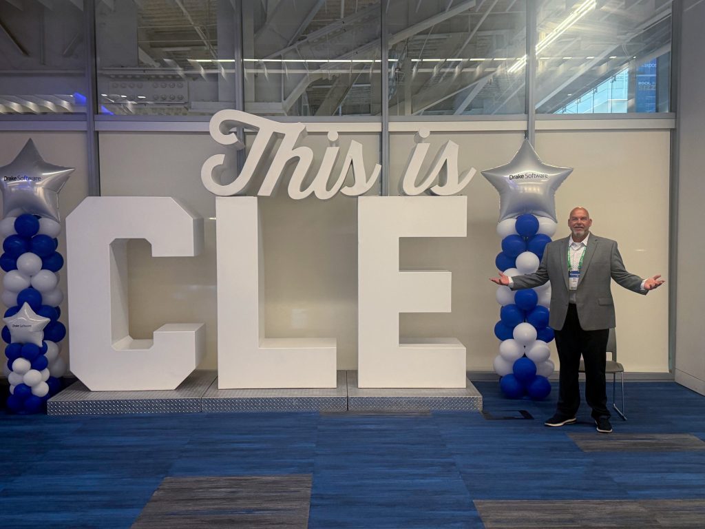 Mark, a TaxBandits Partnerships Manager, standing beside a large "CLE" display at the NATP TaxPosium conference, in Cleveland, Ohio; highlighting professional networking and continuing education for tax professionals.